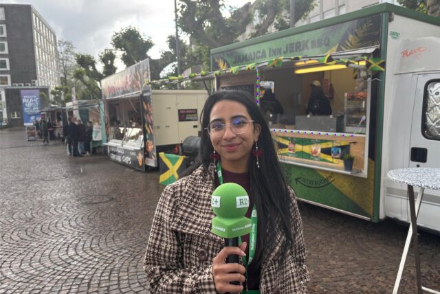 Rosi Taslima Azam steht mit grünem Mikrofon auf dem Rathausplatz in Recklinghausen. Dahinter sind Food-Trucks zu sehen.