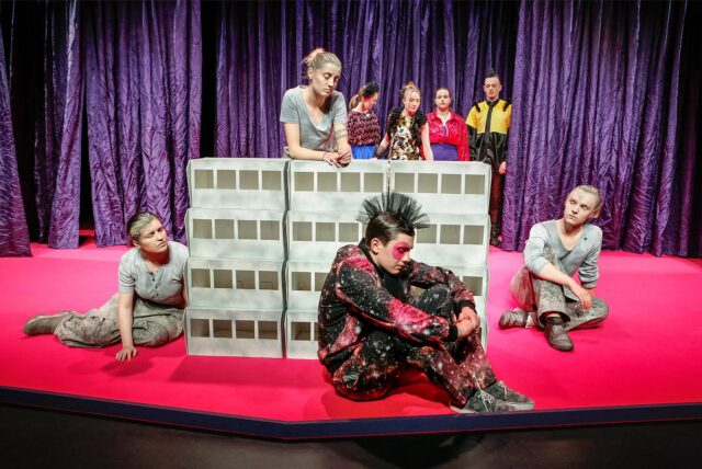 Schauspielerinnen und Schauspieler des Stücks „Betonklotz 2000“ um eine Hochhausblock-Attrappe positioniert.