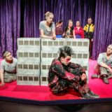 Schauspielerinnen und Schauspieler des Stücks „Betonklotz 2000“ um eine Hochhausblock-Attrappe positioniert.