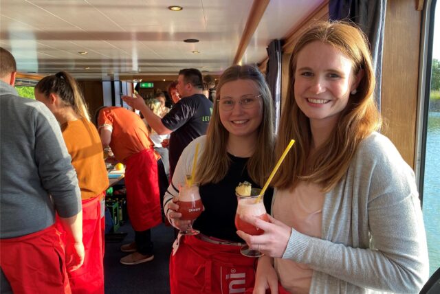 Zwei Frauen mit einem Cocktail in der Hand auf einem Schiff. Sie schauen in die Kamera und lachen.