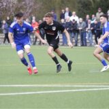 Drei Fußballer kämpfen um einen Ball, im Hintergrund sind Zuschauer auf einer kleinen Tribüne zu sehen.
