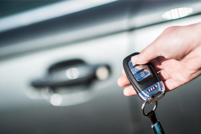 Eine Person hält einen Autoschlüssel in die Kamera. Im Hintergrund erkennt man die Tür eines Autos