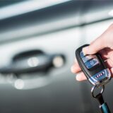 Eine Person hält einen Autoschlüssel in die Kamera. Im Hintergrund erkennt man die Tür eines Autos