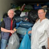 Zwei Personen stehen vor einer Garage, die mit Säcken unterschiedlicher Größe und Farbe gefüllt ist. Die Säcke scheinen mit Kleidung oder Textilien gefüllt zu sein.