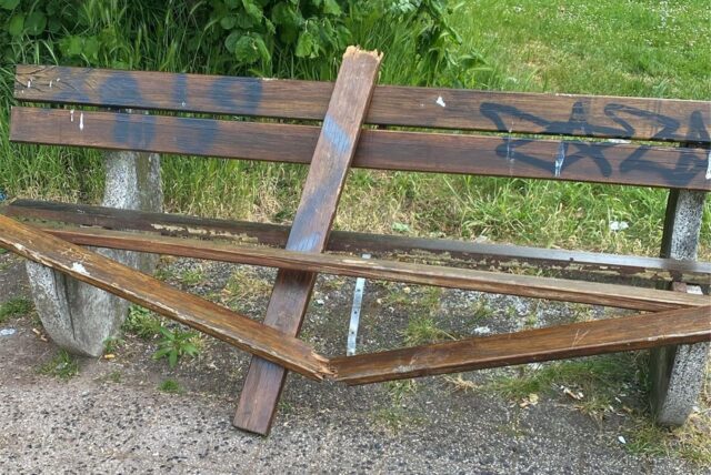 Eine beschädigte Parkbank steht auf einem befestigten Weg. Die Bank ist teilweise zerbrochen und mit Graffiti beschmiert.