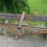 Eine beschädigte Parkbank steht auf einem befestigten Weg. Die Bank ist teilweise zerbrochen und mit Graffiti beschmiert.