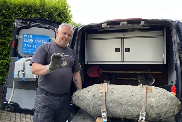 Ein Mann steht an einer alten Weltkriegsbombe und hält einen Zünder in der Hand.