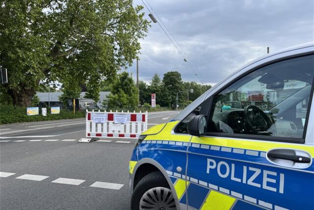Ein Polizei-Fahrzeug steht am Rande einer Straße, die durch eine rot-weiß gestreifte Barriere blockiert wird. Schilder auf der Barriere weisen auf Bauarbeiten oder eine Straßensperrung hin.