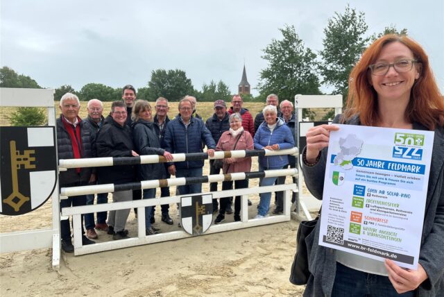 Bianca Gawollek von der Familienbildungsstätte zeigt ein Plakat, im Hintergrund eine Menschengruppe hinter dem Parcours-Hindernis mit Stadtwappen.