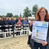 Bianca Gawollek von der Familienbildungsstätte zeigt ein Plakat, im Hintergrund eine Menschengruppe hinter dem Parcours-Hindernis mit Stadtwappen.