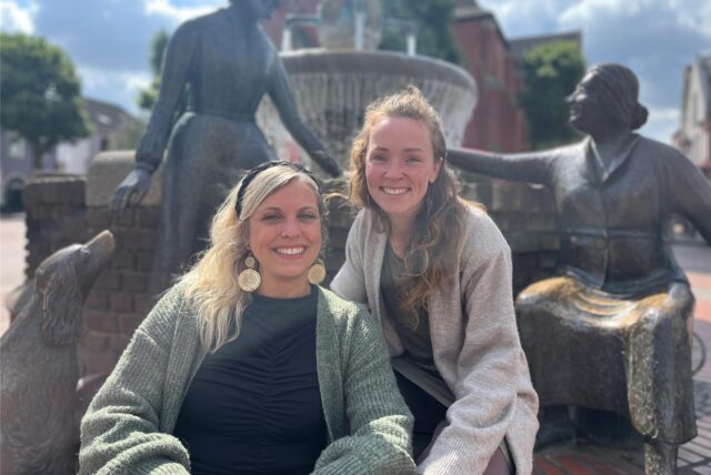 Jasmin Roß und Rahel Artmann sitzen am Brunnen am Marktplatz in Haltern am See und lächeln in die Kamera. Hinter ihnen sind die