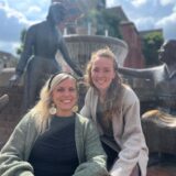 Jasmin Roß und Rahel Artmann sitzen am Brunnen am Marktplatz in Haltern am See und lächeln in die Kamera. Hinter ihnen sind die