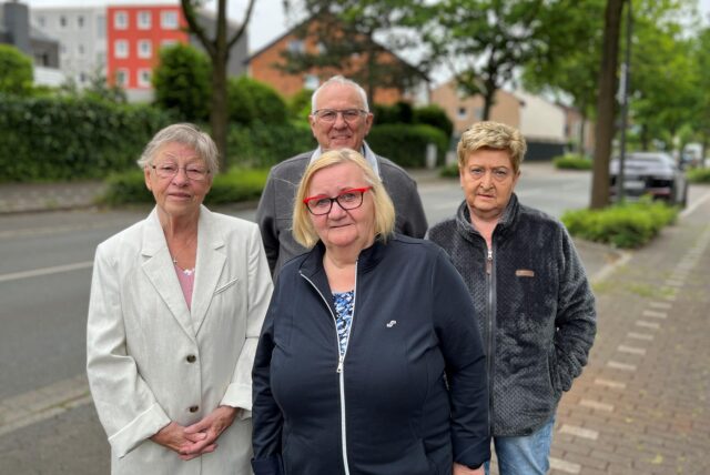Eine Gruppe von vier Personen steht an einem bewölkten Tag auf einem Gehweg neben einer Straße. Im Hintergrund sind Wohnhäuser und Bäume zu sehen.