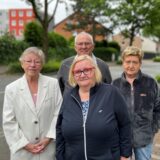 Eine Gruppe von vier Personen steht an einem bewölkten Tag auf einem Gehweg neben einer Straße. Im Hintergrund sind Wohnhäuser und Bäume zu sehen.