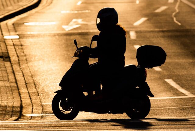 Rollerfahrer auf Roller im Gegenlicht erscheint schwarz vor einer helleren Straße mit Fahrbahnmarkierungen.