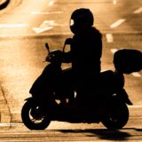 Rollerfahrer auf Roller im Gegenlicht erscheint schwarz vor einer helleren Straße mit Fahrbahnmarkierungen.