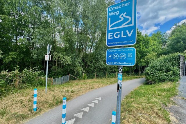 Das Bild zeigt einen Abschnitt des Emscherwegs, einem Fußgänger- und Fahrradweg, gekennzeichnet durch blaue Schilder der Emschergenossenschaft EGLV. Ein Schild weist darauf hin, dass auf dem Weg kein Winterdienst erfolgt.