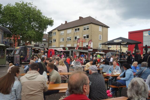 Viele Menschen sitzen auf Bänken, dahinter stehen Foodtrucks.