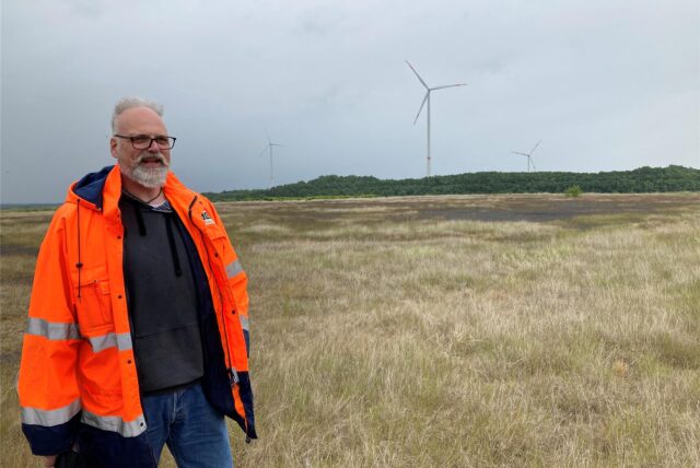 RAG-Geologe Oliver Kunz steht auf einer Halde vor einem Windrad.