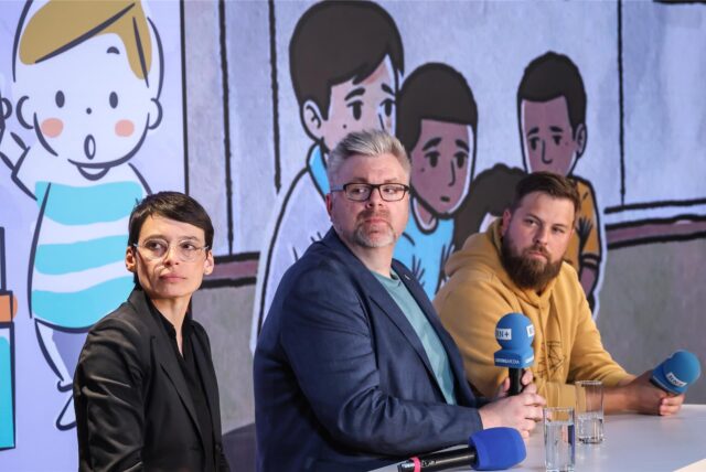 NRW-Familienministerin Josefine Paul und zwei Männer stzen auf einem Podium und schauen einen Film an. Im Hintergrund zu sehen sind Zeichnungen von unglücklichen Kindern.