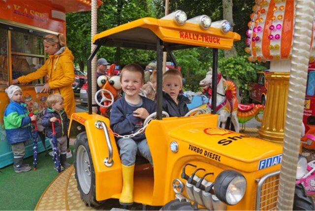 Zwei Kinder sitzen in einem gelben Traktor auf einem Karussell. Im Hintergrund ist ein Kassenhäuschen und weitere Kinder zu sehen.