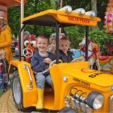 Zwei Kinder sitzen in einem gelben Traktor auf einem Karussell. Im Hintergrund ist ein Kassenhäuschen und weitere Kinder zu sehen.