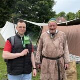 Ein Reporter mit Mikrofon der WZ+ interviewt einen Mann in historischer Kleidung vor Zelten. Im Hintergrund ist ein Schild mit mittelalterlicher Schrift zu sehen.