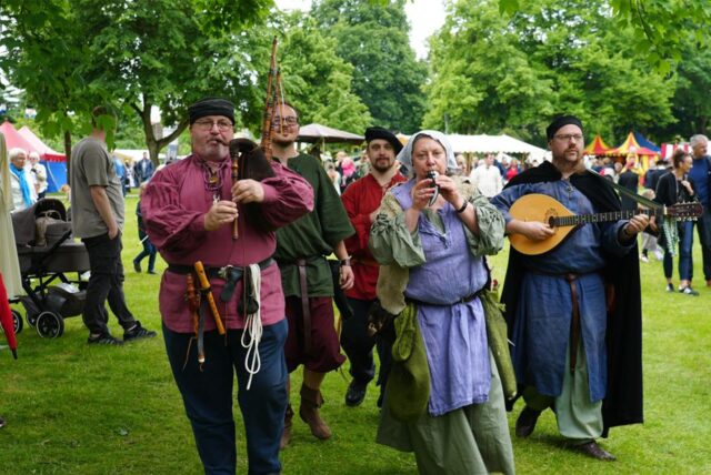 Eine Gruppe von Personen in historisch anmutenden Kostümen spielt auf mittelalterlichen Musikinstrumenten in einem Park.  Im Hintergrund sind Menschen und Zelte zu sehen, was auf ein Festival oder einen Markt hindeutet.