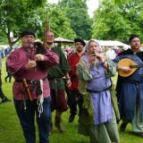 Eine Gruppe von Personen in historisch anmutenden Kostümen spielt auf mittelalterlichen Musikinstrumenten in einem Park.  Im Hintergrund sind Menschen und Zelte zu sehen, was auf ein Festival oder einen Markt hindeutet.