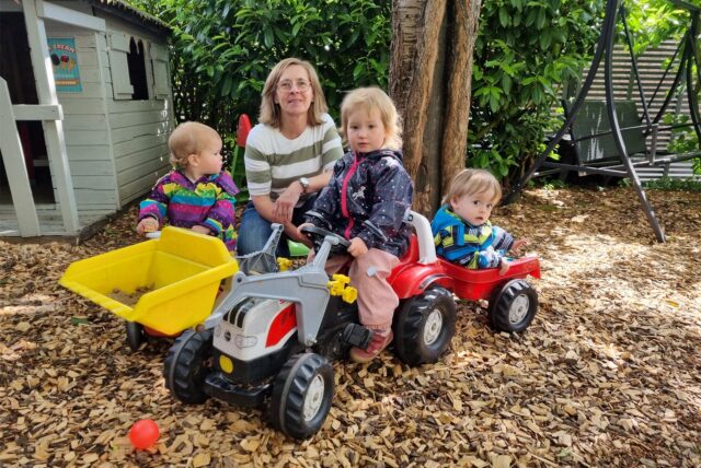 Drei Kinder spielen vor einer hockenden Frau mit einem Spielzeugtrecker und einem Bobbycar.
