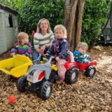 Drei Kinder spielen vor einer hockenden Frau mit einem Spielzeugtrecker und einem Bobbycar.