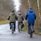 Drei Personen fahren auf Fahrrädern einen Weg entlang, umgeben von Bäumen und Büschen. Die Radfahrer tragen Jacken und sind in Bewegung.