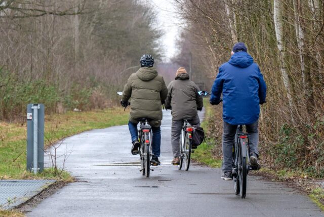 Drei Personen fahren auf Fahrrädern einen Weg entlang, umgeben von Bäumen und Büschen. Die Radfahrer tragen Jacken und sind in Bewegung.