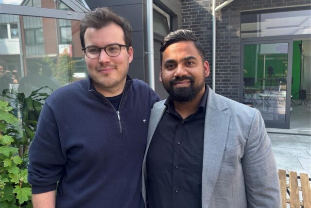 Zwei Männer schütteln sich die Hände vor einem Gebäude. Der Mann links trägt eine dunkle Jacke und beige Hosen, während der Mann rechts ein graues Sakko über einem schwarzen Hemd und schwarze Hosen trägt.