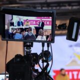 Auf dem kleinen Bildschirm an einer professionellen Videokamera sind mehrere Menschen auf einem Podium zu sehen.