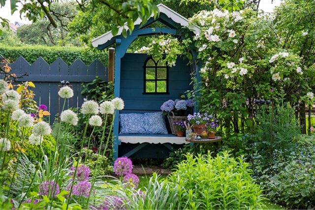 Offene Gartenpforte 2025: Blick in einen blühenden Garten mit einer überdachten blauen Gartenbank inmitten von Staudenbeeten.