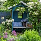 Offene Gartenpforte 2025: Blick in einen blühenden Garten mit einer überdachten blauen Gartenbank inmitten von Staudenbeeten.