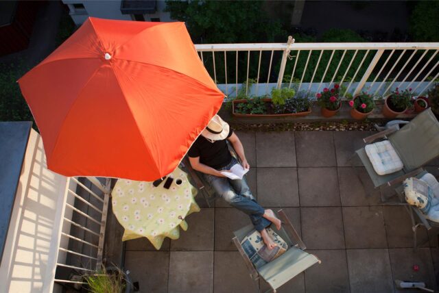 Ein Mann sitzt auf dem Balkon unter einem Sonnenschirm und liest ein Buch.
