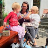 Eine Tagesmutter aus Dortmund redet bei Sonnenschein mit zwei Kindern – um sie herum liegen ein paar Spielsachen.