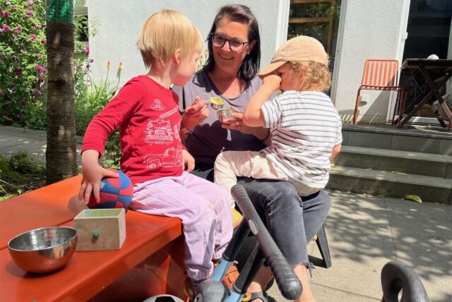 Eine Tagesmutter aus Dortmund redet bei Sonnenschein mit zwei Kindern – um sie herum liegen ein paar Spielsachen.