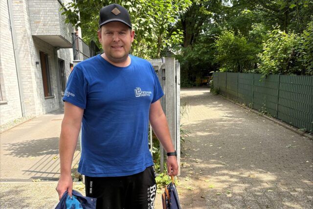 Ein Mann steht im Freien, bekleidet mit einem blauen T-Shirt  und schwarzen Shorts mit dem. Er trägt eine dunkle Kappe und hält eine Jacke sowie eine Tasche in den Händen.