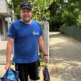 Ein Mann steht im Freien, bekleidet mit einem blauen T-Shirt  und schwarzen Shorts mit dem. Er trägt eine dunkle Kappe und hält eine Jacke sowie eine Tasche in den Händen.
