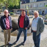 Drei Männer stehen auf einem Platz vor einem Brunnen.