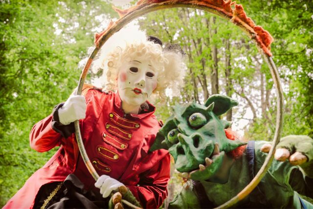 Eine Frau in Kostüm und mit Maske sowie eine Drachenfigur vom Straßentheater „Unbedacht“, das beim Kunstmarkt 2025 in Herten auftritt, schauen durch einen Hula-Hoop-Reifen.