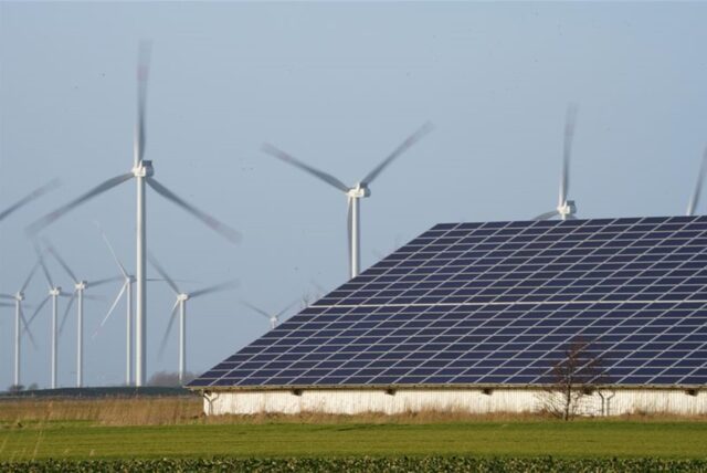 Windenergieanlagen stehen neben einer Halle mit Photovoltaik-Anlagen auf dem Dach.