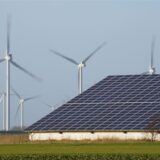 Windenergieanlagen stehen neben einer Halle mit Photovoltaik-Anlagen auf dem Dach.