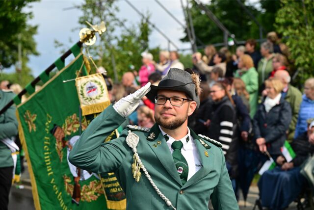 Ein Mann in Uniform und Hut salutiert bei einer Veranstaltung im Freien. Im Hintergrund befinden sich Zuschauer und eine Fahne des Heimatvereins.