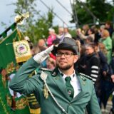 Ein Mann in Uniform und Hut salutiert bei einer Veranstaltung im Freien. Im Hintergrund befinden sich Zuschauer und eine Fahne des Heimatvereins.