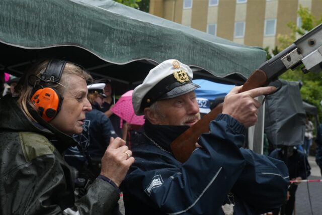 Ein Mann in Marineuniform zielt mit einem Gewehr, während eine Frau mit Gehörschutz danebensteht. Die Szene spielt sich im Freien unter einem Unterstand an einem regnerischen Tag ab.