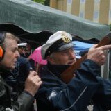 Ein Mann in Marineuniform zielt mit einem Gewehr, während eine Frau mit Gehörschutz danebensteht. Die Szene spielt sich im Freien unter einem Unterstand an einem regnerischen Tag ab.
